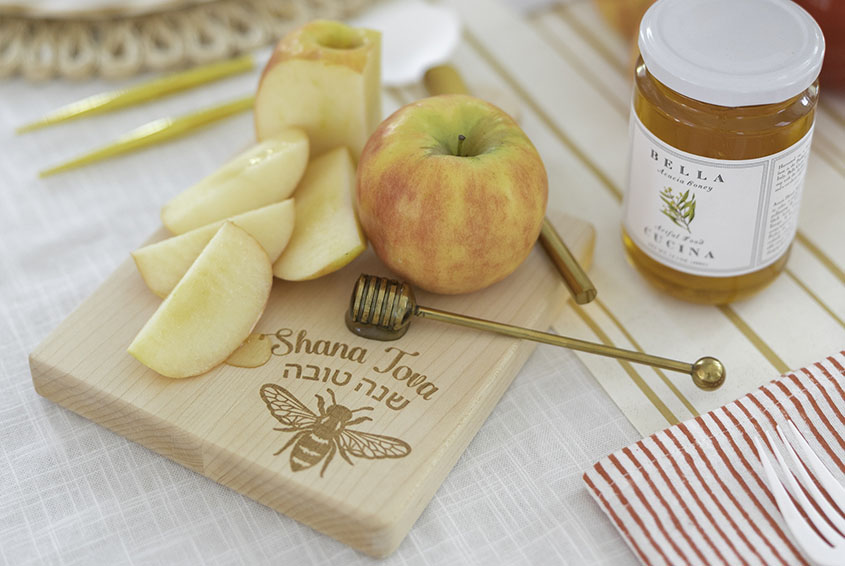 Sliced apple, whole apple, honey jar, and honey dipper on a board with Hebrew text and bee engraving.