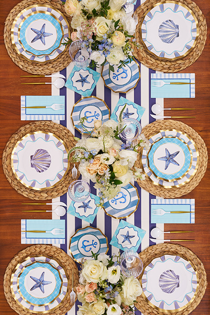An overhead view of a long dining table set with nautical-themed plates, a striped runner, and floral arrangements.