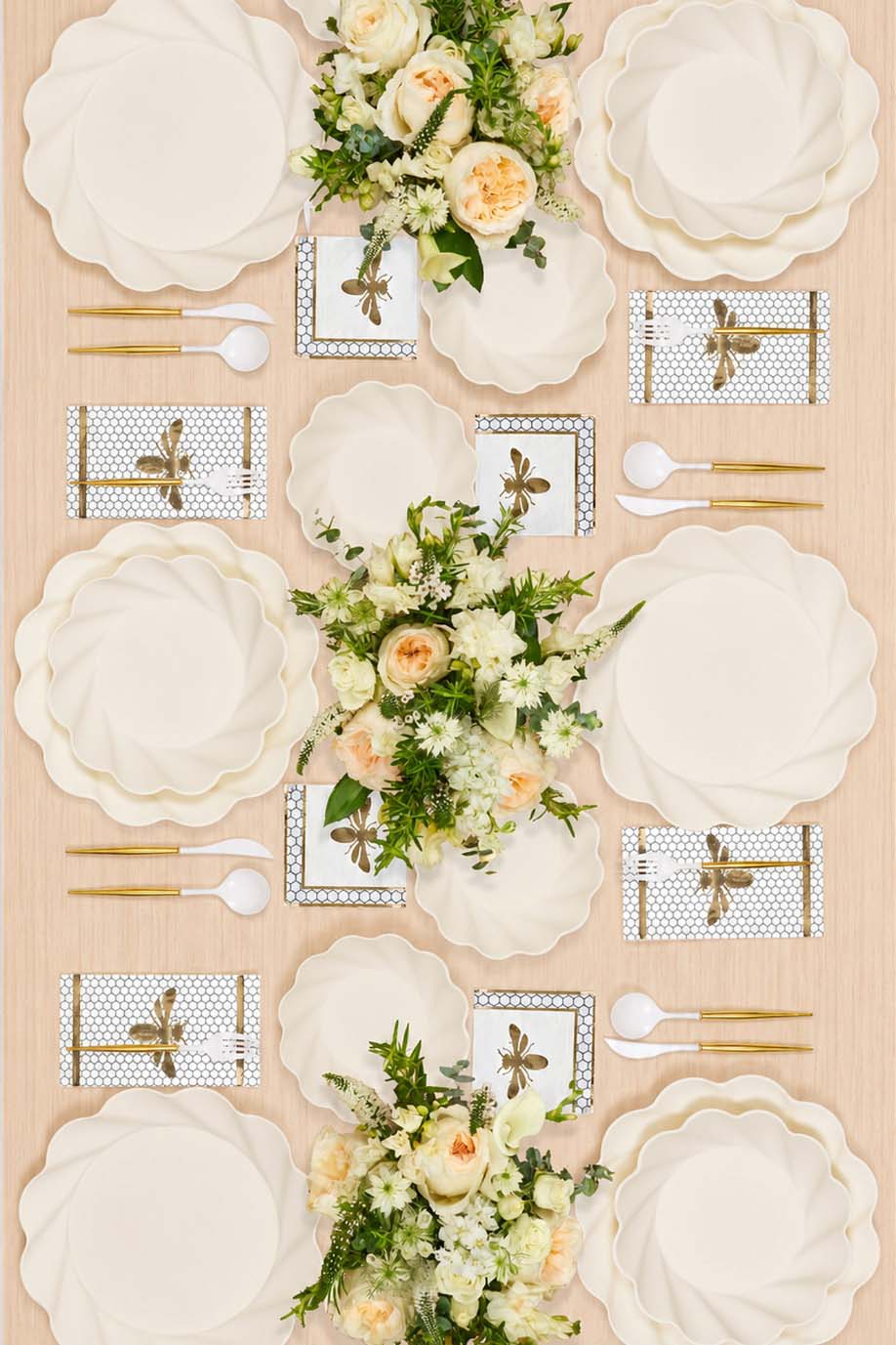 An overhead view of a tablescape with scalloped plates, bee-themed napkins, gold flatware, and a floral centerpiece.