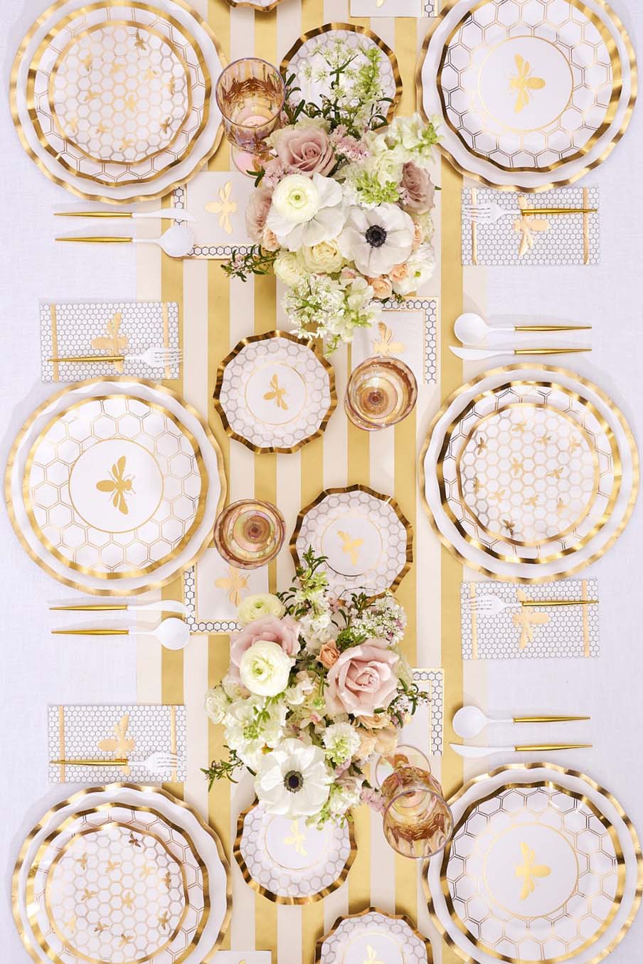 An overhead view of a bee-themed tablescape with gold and white plates, gold cutlery, and floral centerpieces.