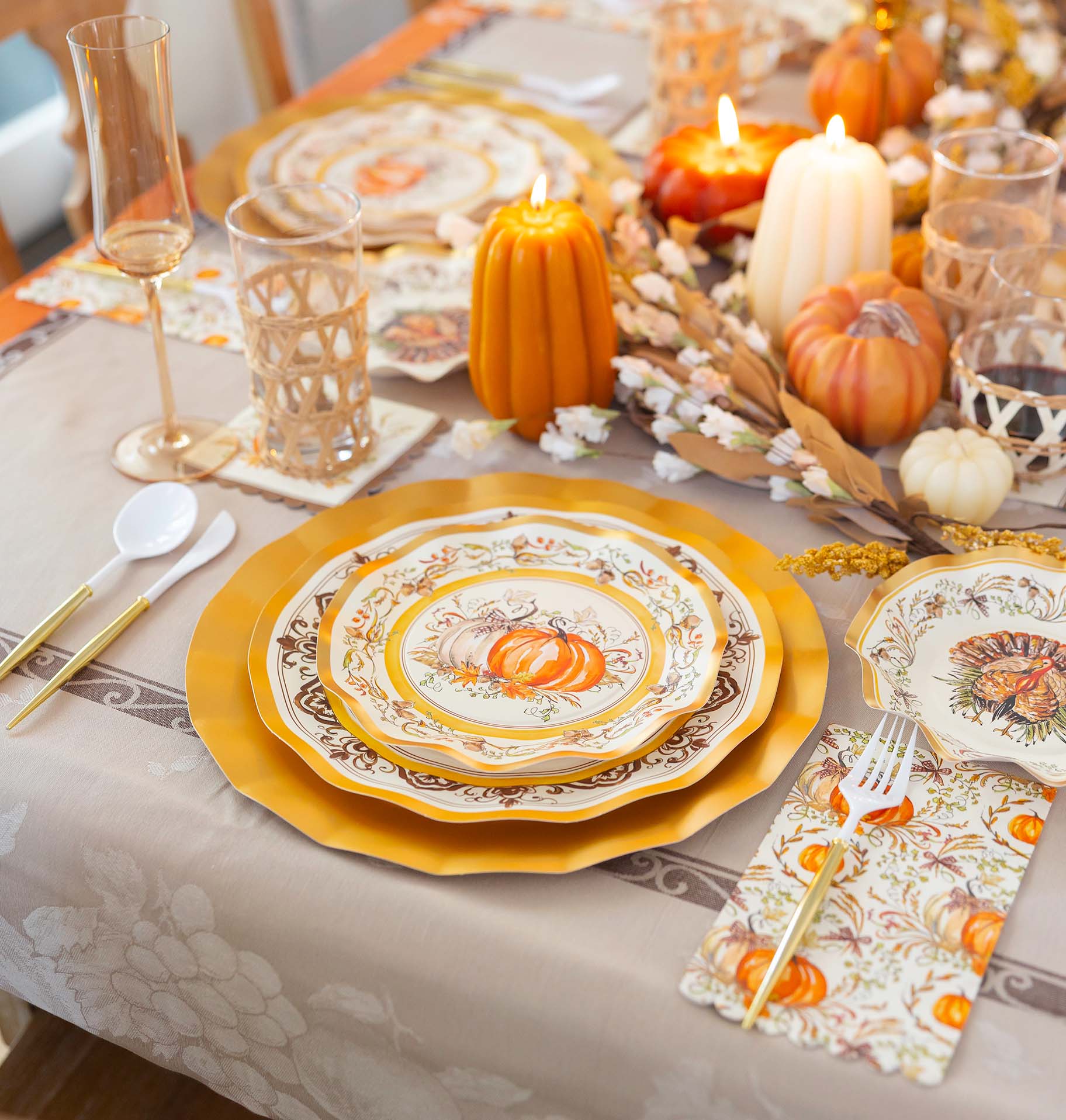 A Thanksgiving table setting with pumpkin-themed plates, gold chargers, decorative candles, and plastic cutlery.