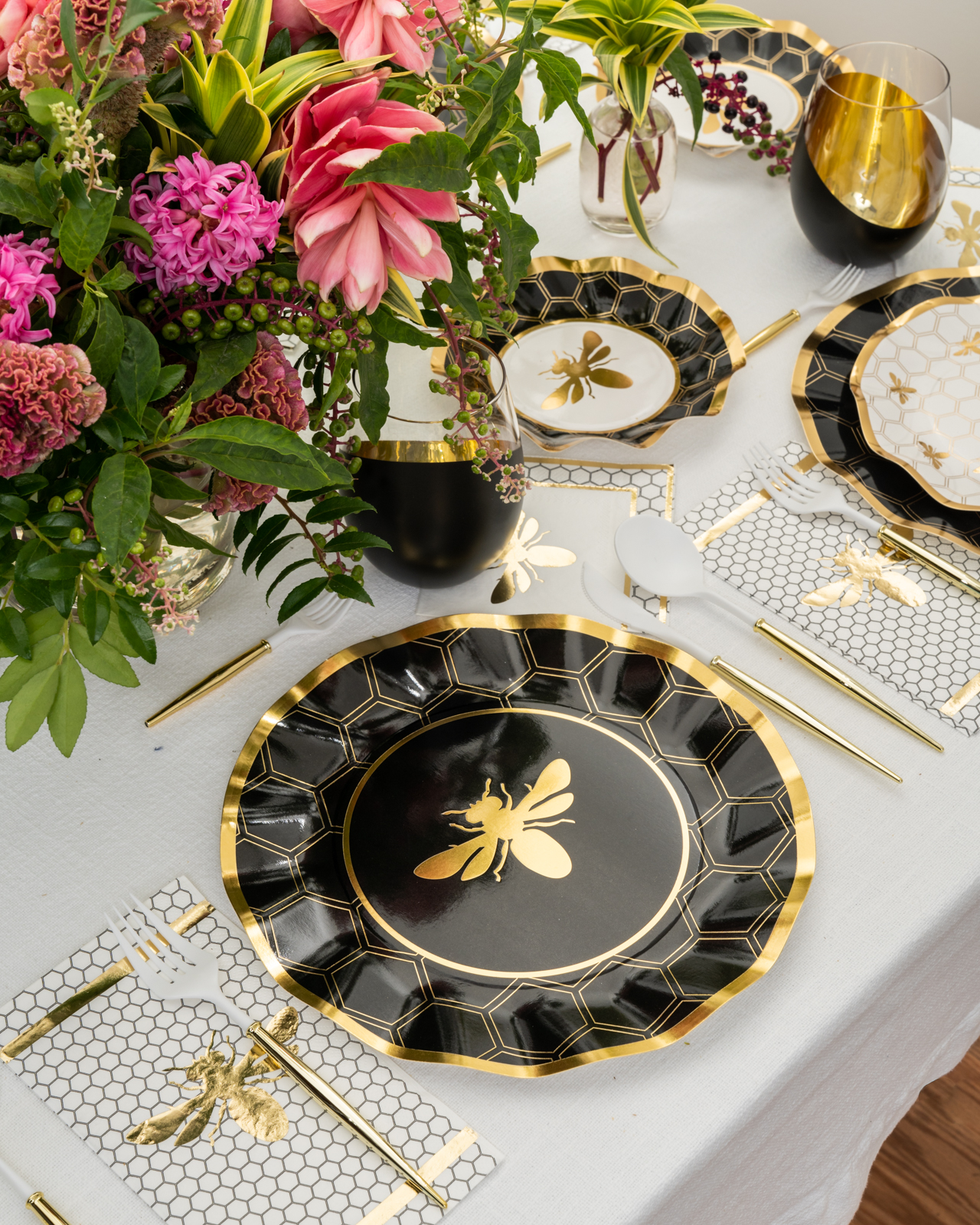 A bee-themed table setting with black and gold paper plates and a large pink floral arrangement.