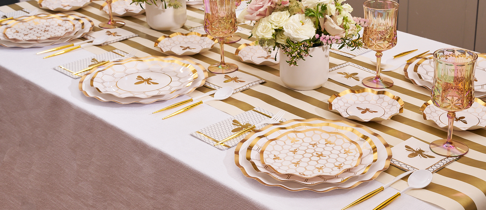 A beautifully set table with bee-themed plates, gold cutlery, iridescent glasses, and a floral centerpiece.