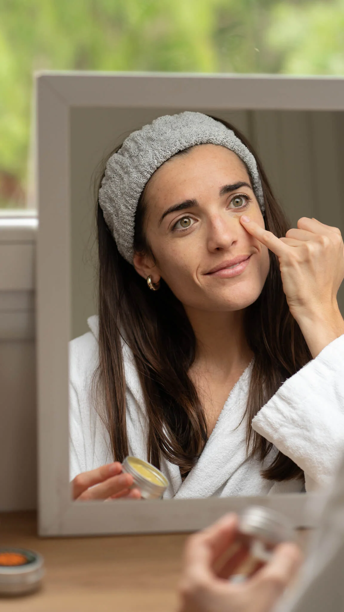Woman in a robe applying skincare cream in front of a mirror.