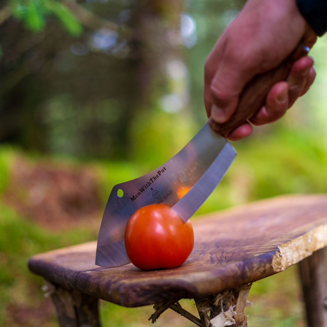 Hand holding a knife cutting a tomato on a wooden surface outdoors.