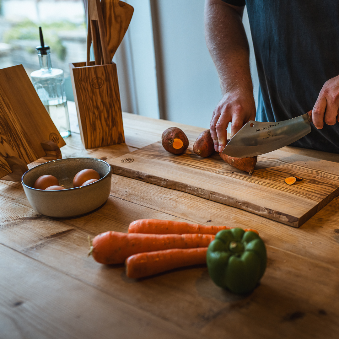 Person chopping sweet potatoes on a wooden board with vegetables nearby.