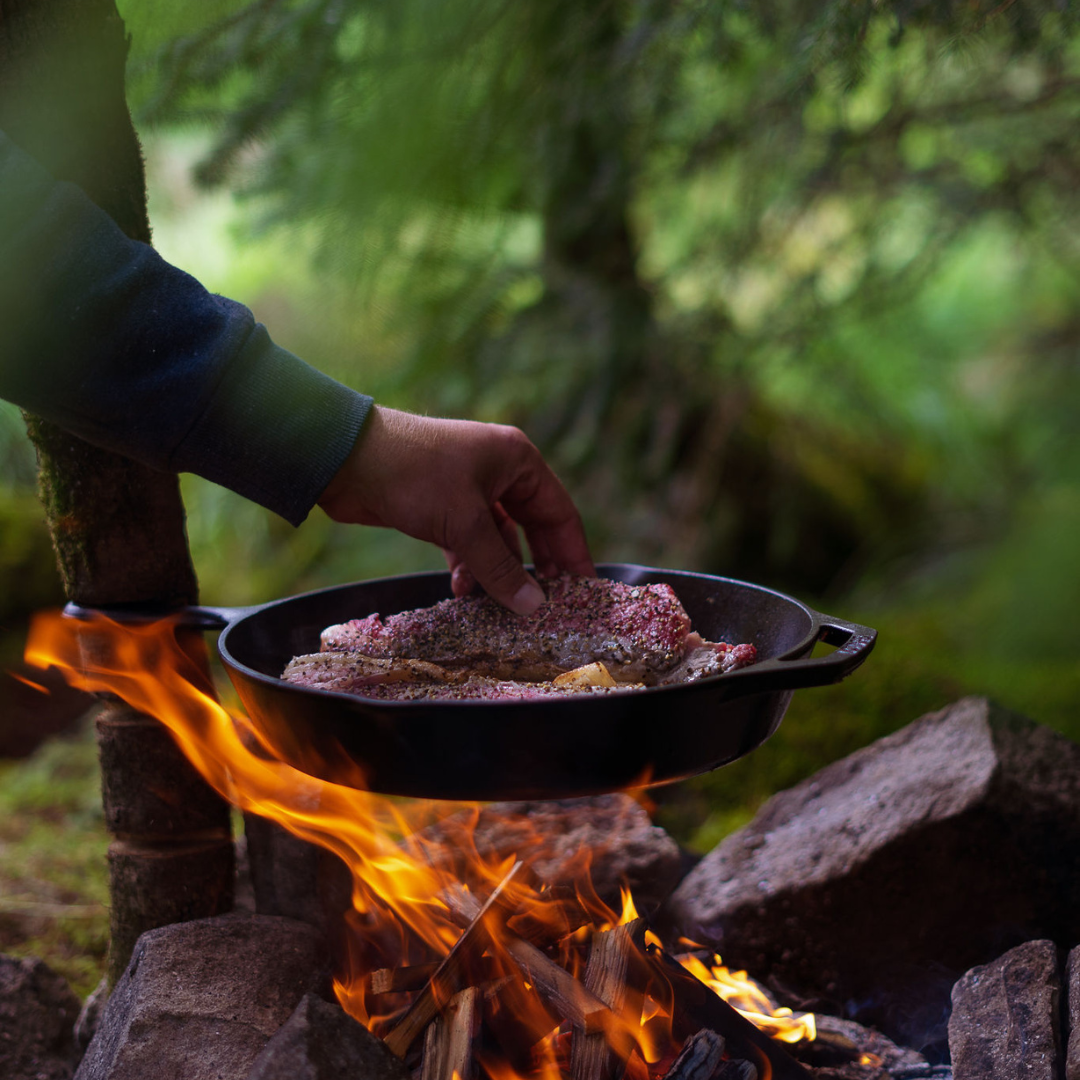 Person cooking meat in a pan over an open fire outdoors.
