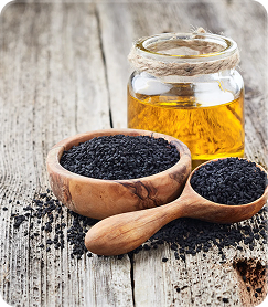 A glass jar of oil sits on a wooden surface next to a wooden bowl and spoon of black seeds.