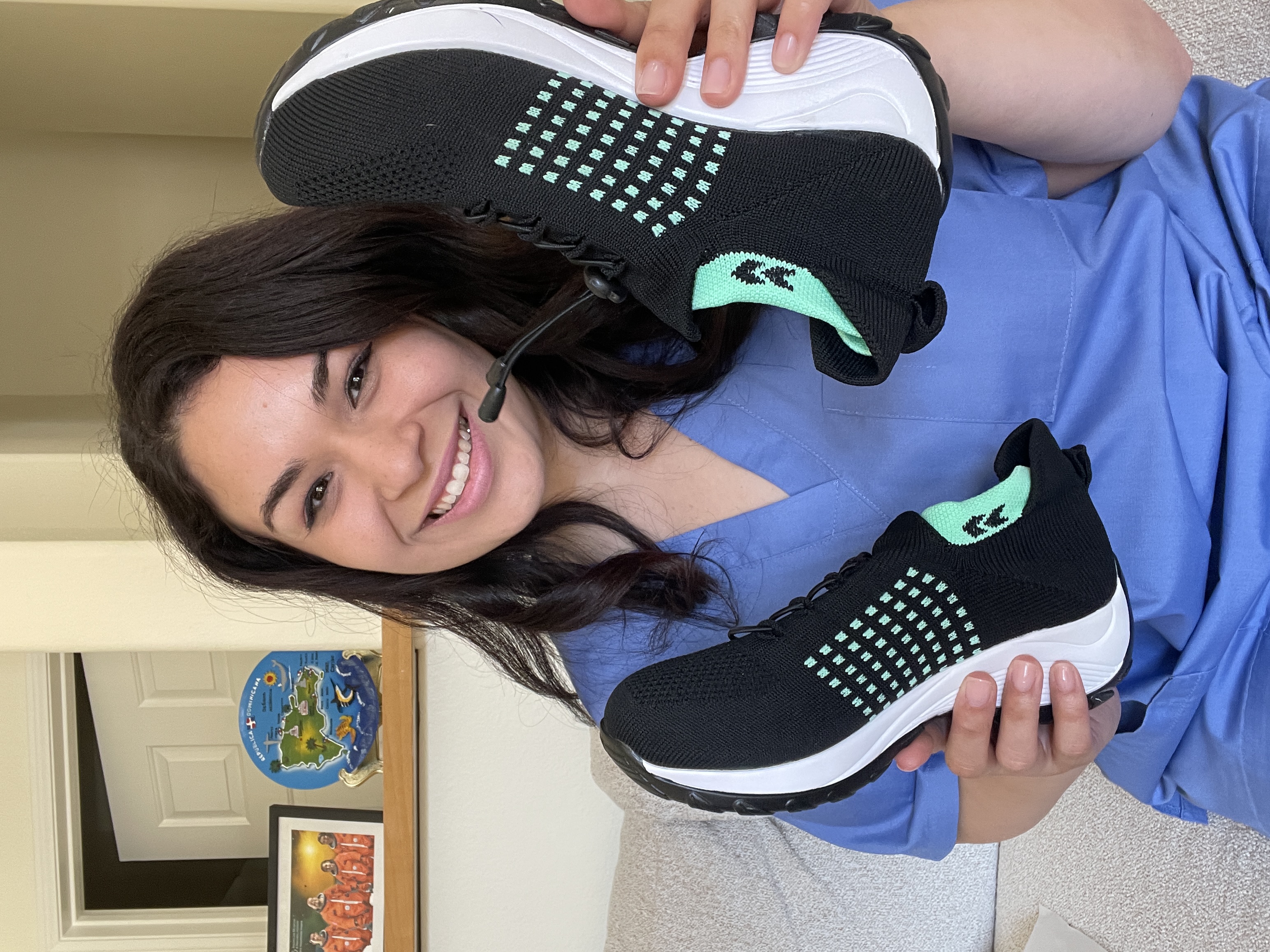 Person in blue scrubs holding a pair of black and green sneakers, smiling.