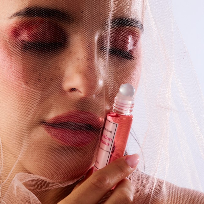 Person with dramatic makeup holding a pink roller bottle, partially covered by a veil.
