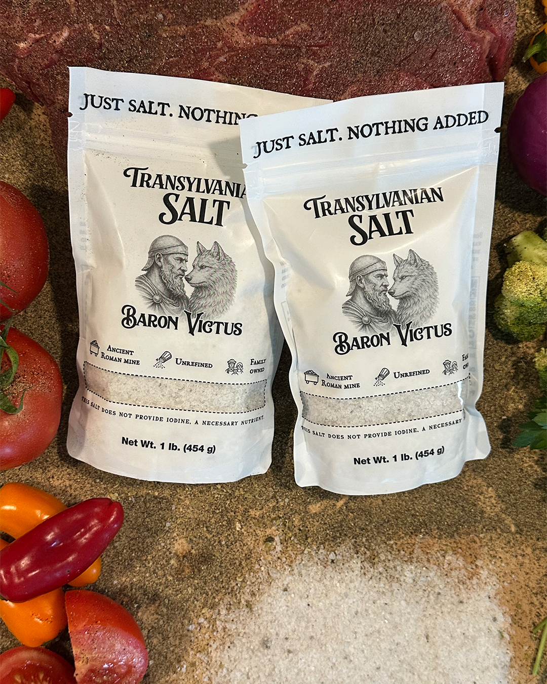 Two pouches of Baron Victus Transylvanian Salt displayed with raw steak, tomatoes, and peppers on a counter.