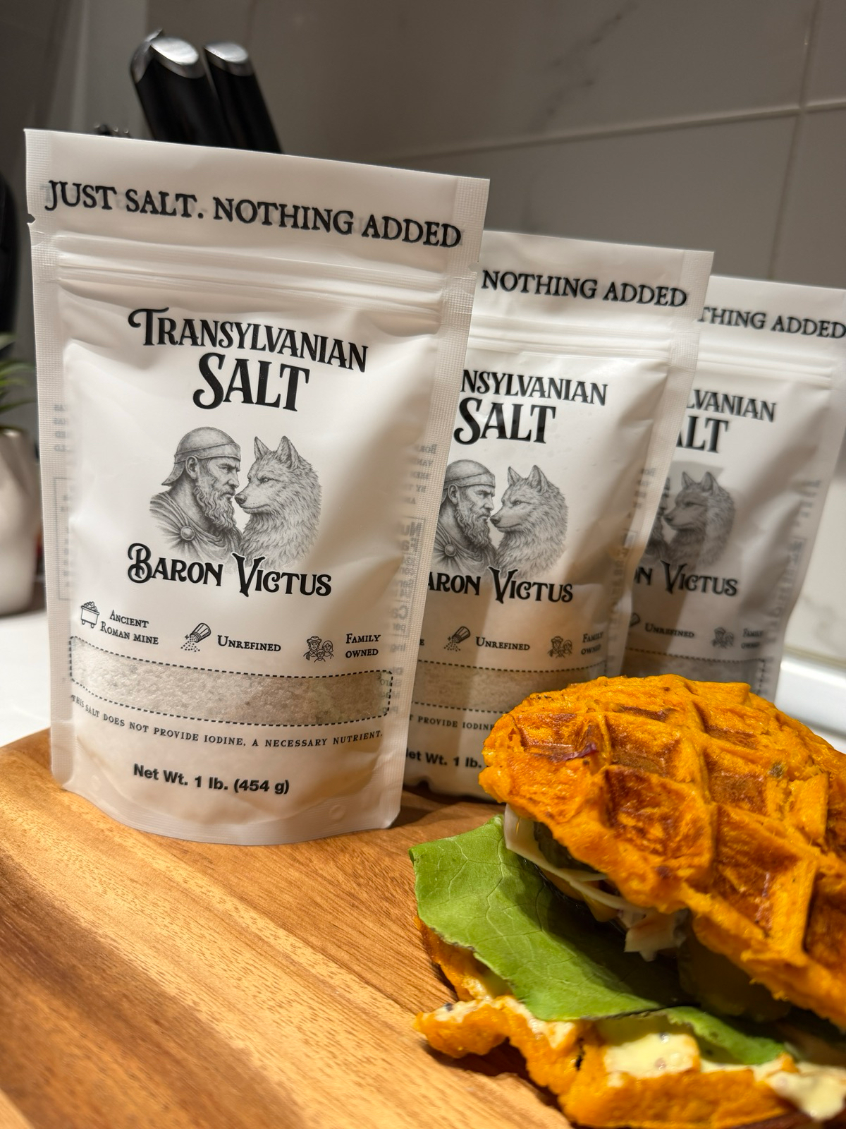 Three bags of Transylvanian Salt arranged on a wooden board behind a waffle sandwich.