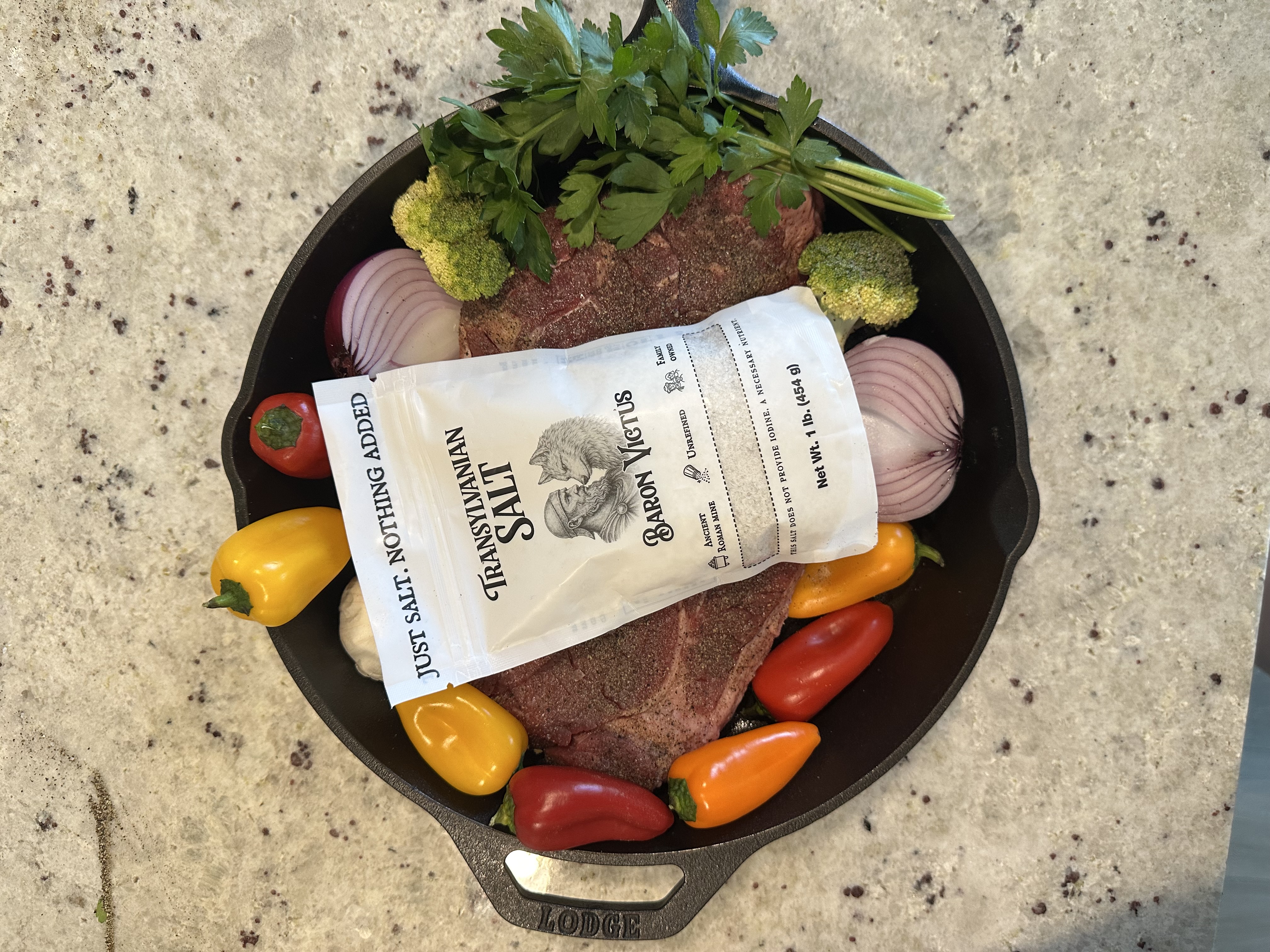 A bag of Transylvanian Salt on a raw steak with fresh vegetables in a cast iron skillet.