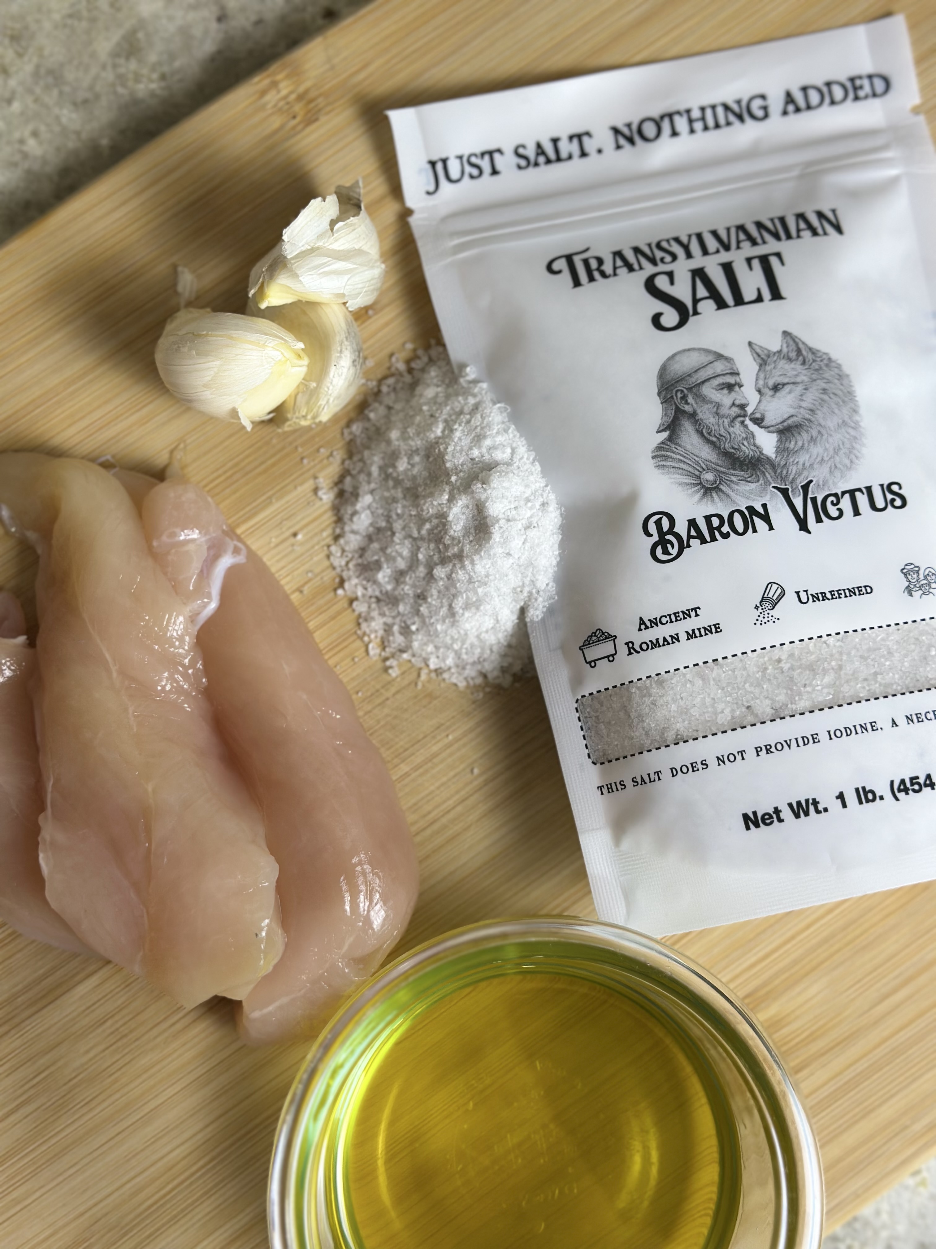 Raw chicken, garlic, salt, and a bowl of oil on a wooden cutting board.