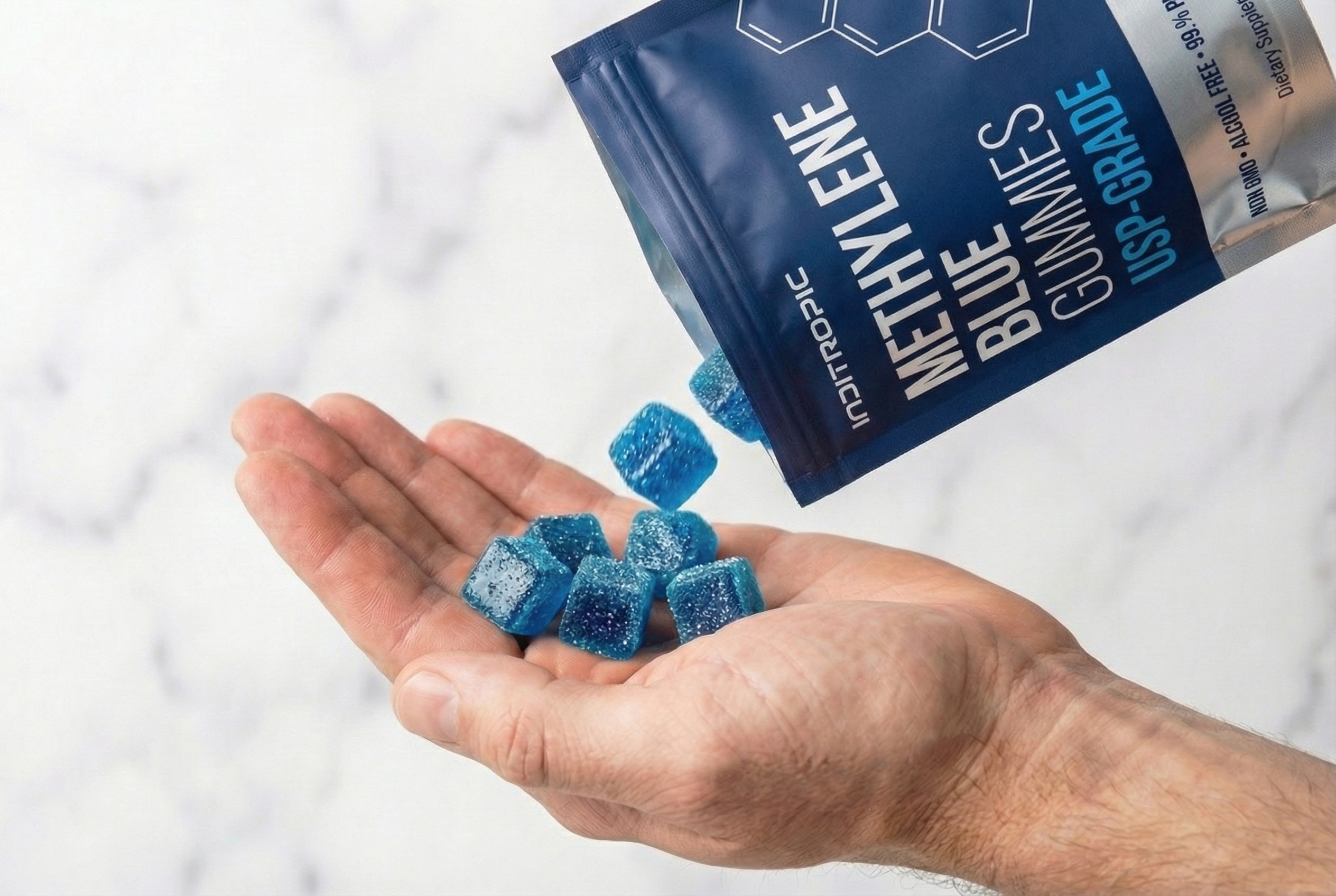 A person pouring blue, cube-shaped gummies from a pouch into their cupped hand against a white marble background.