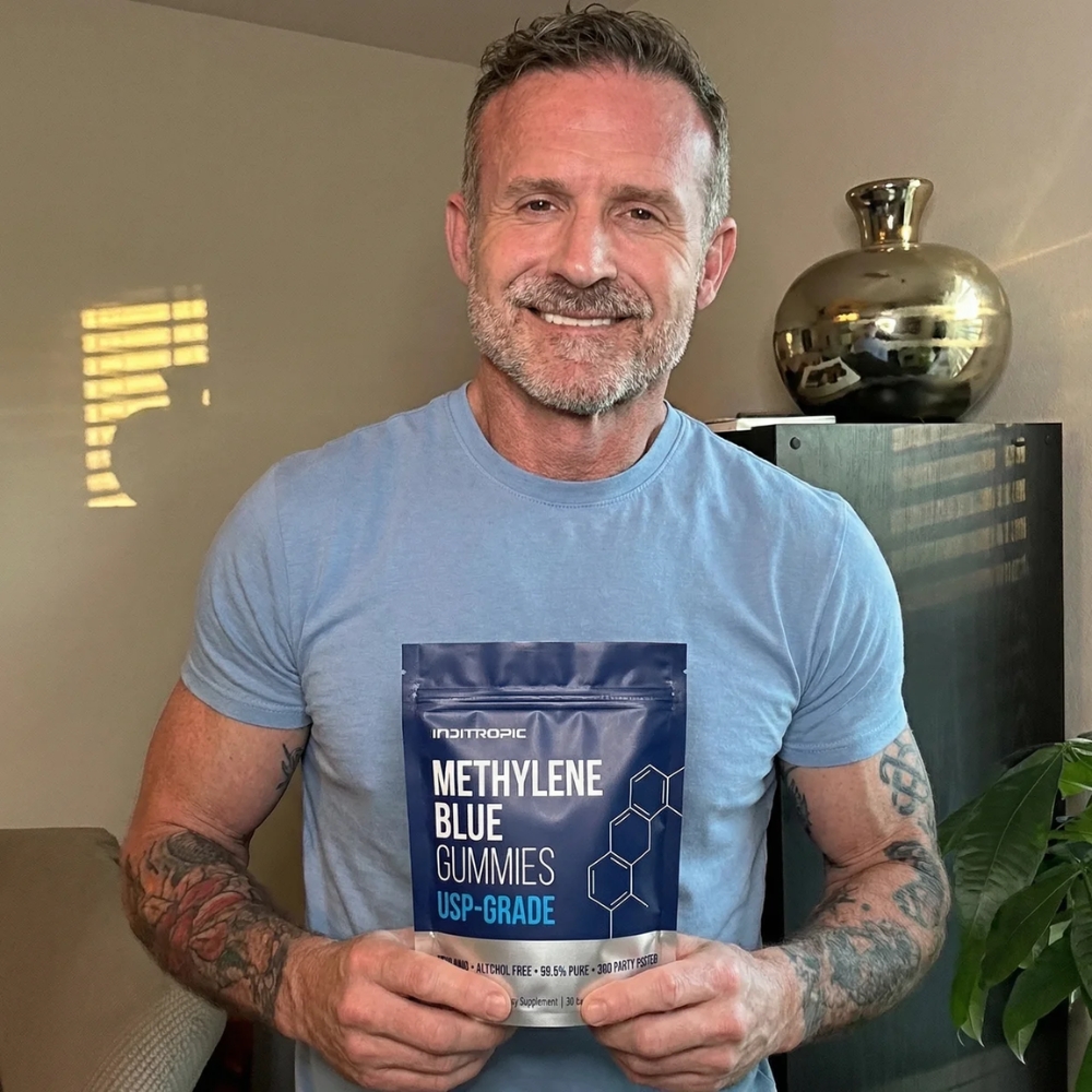 A smiling man with tattoos on his arms holds up a blue pouch of Methylene Blue Gummies.