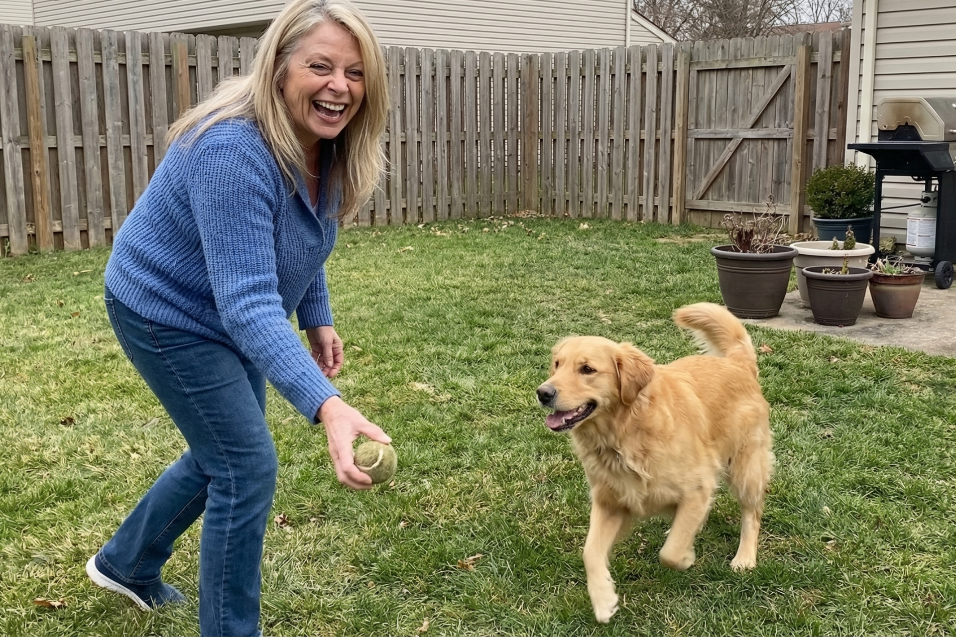 A woman in a blue sweater laughs while playing fetch with a golden retriever in a backyard.