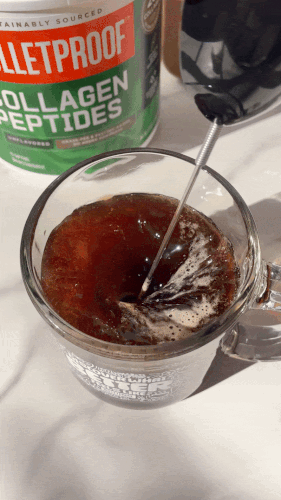 Stirring coffee in a glass mug with a metal frother, collagen peptides container in background.
