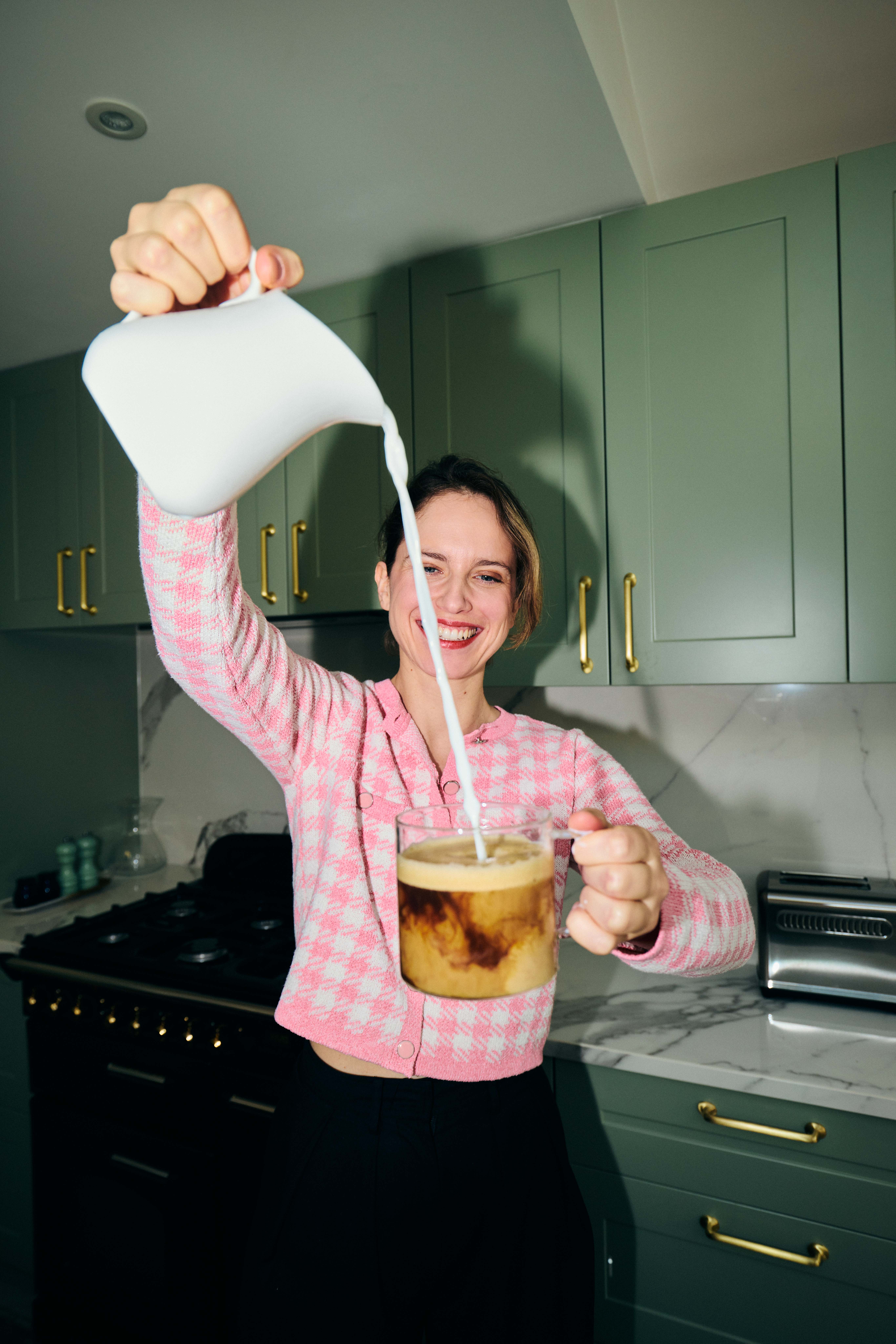 Person pouring milk into a glass of coffee in a kitchen.