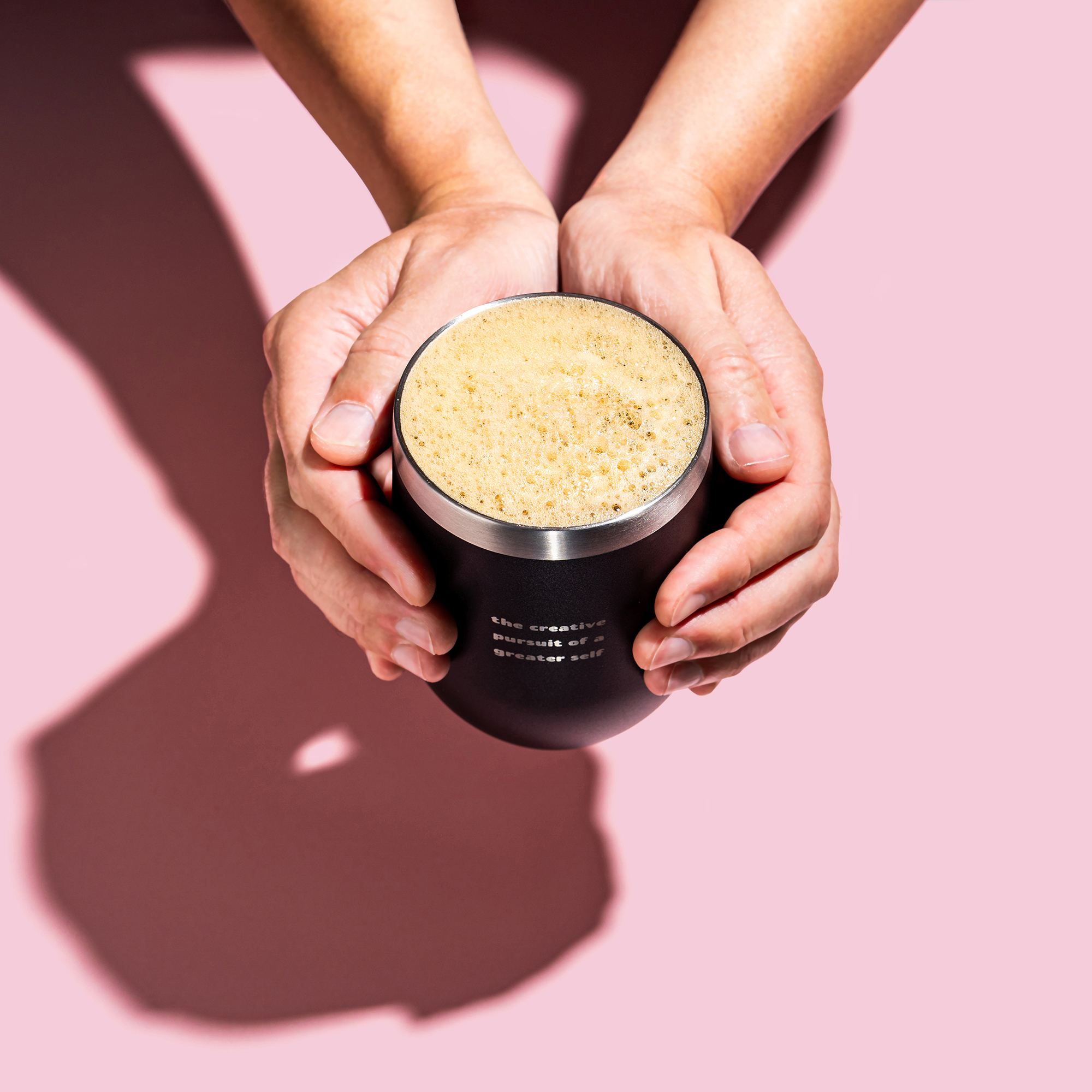 Hands holding a frothy drink in a black cup on pink background.