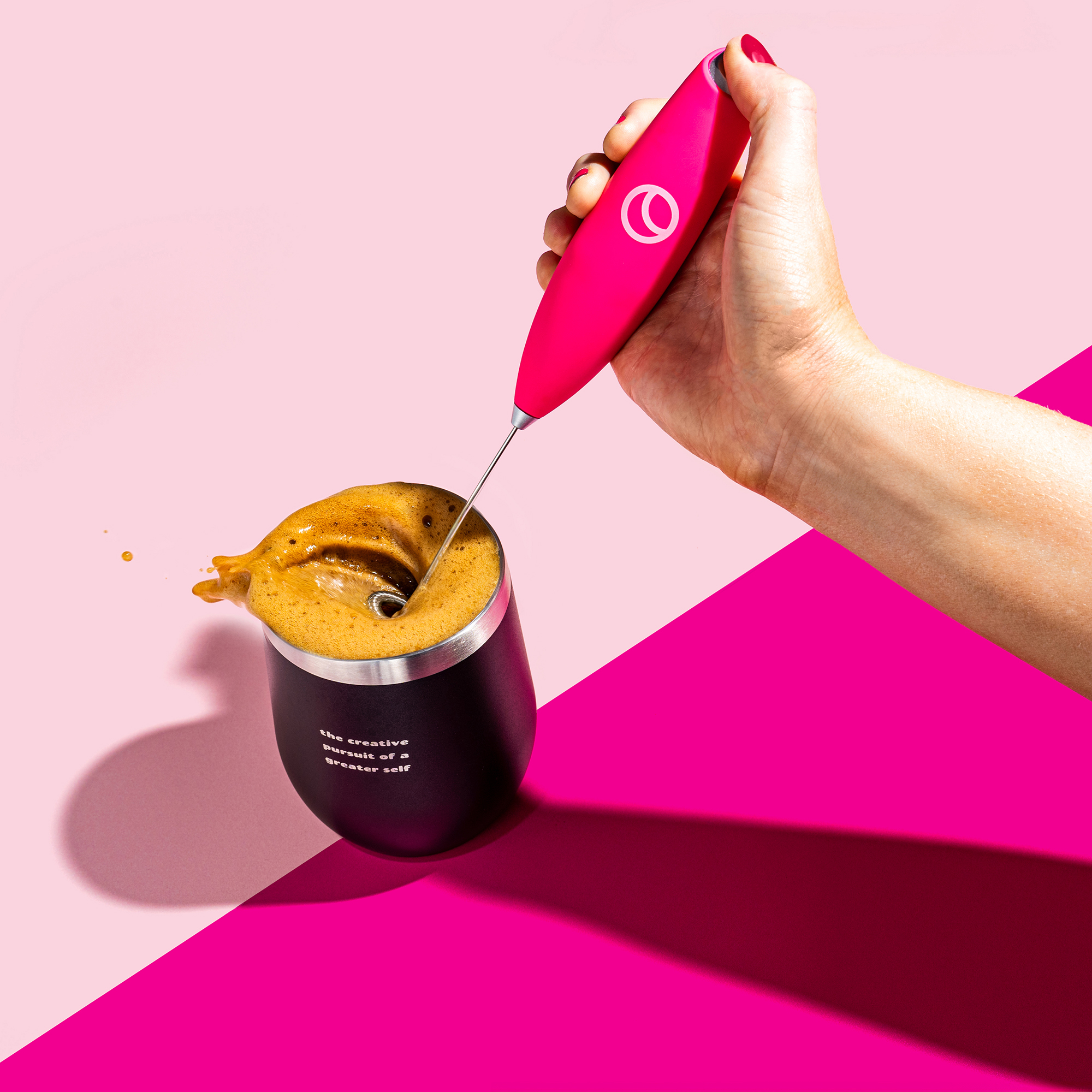 Hand frothing coffee in a black cup with pink background.