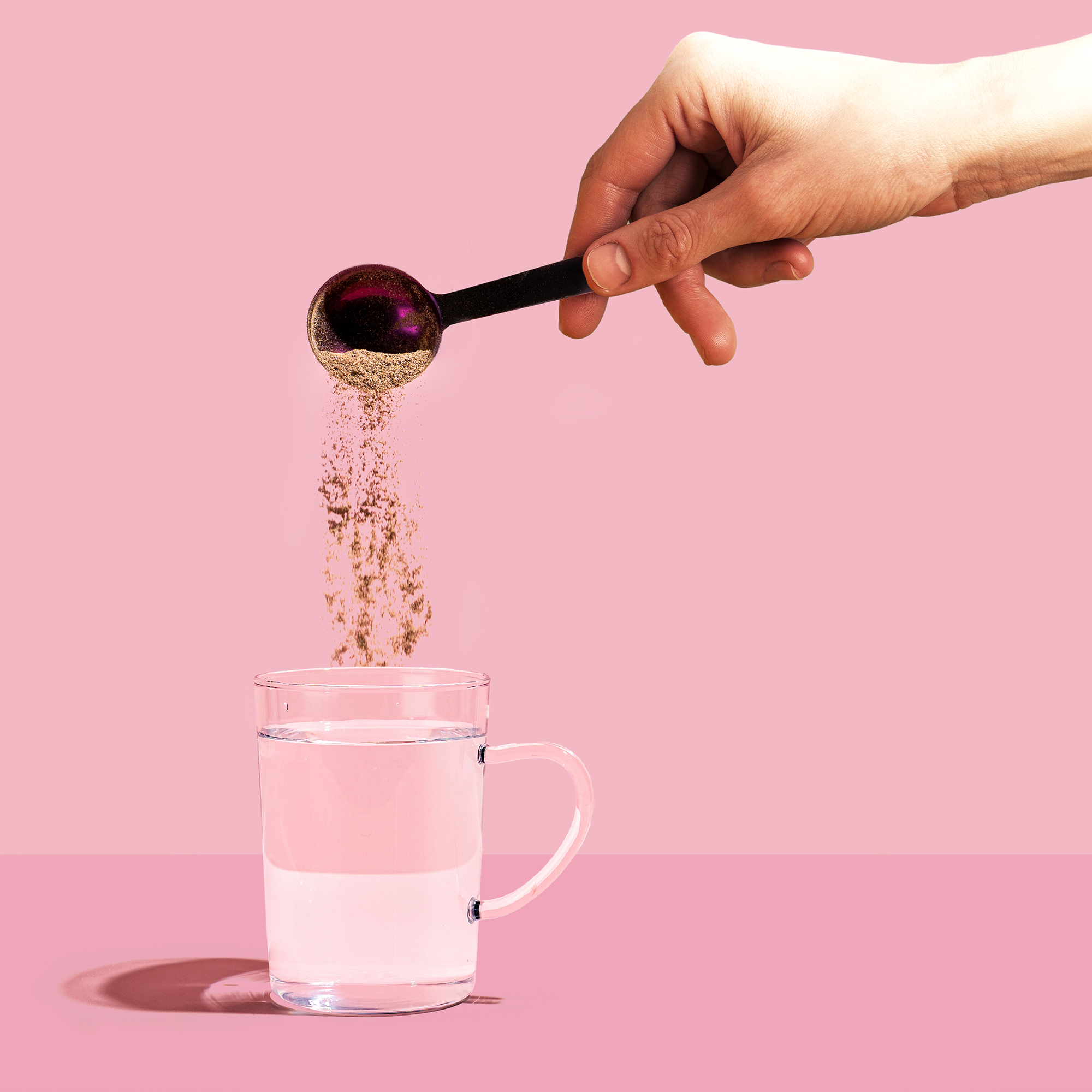 A hand pouring powder from a spoon into a glass of water on pink background.