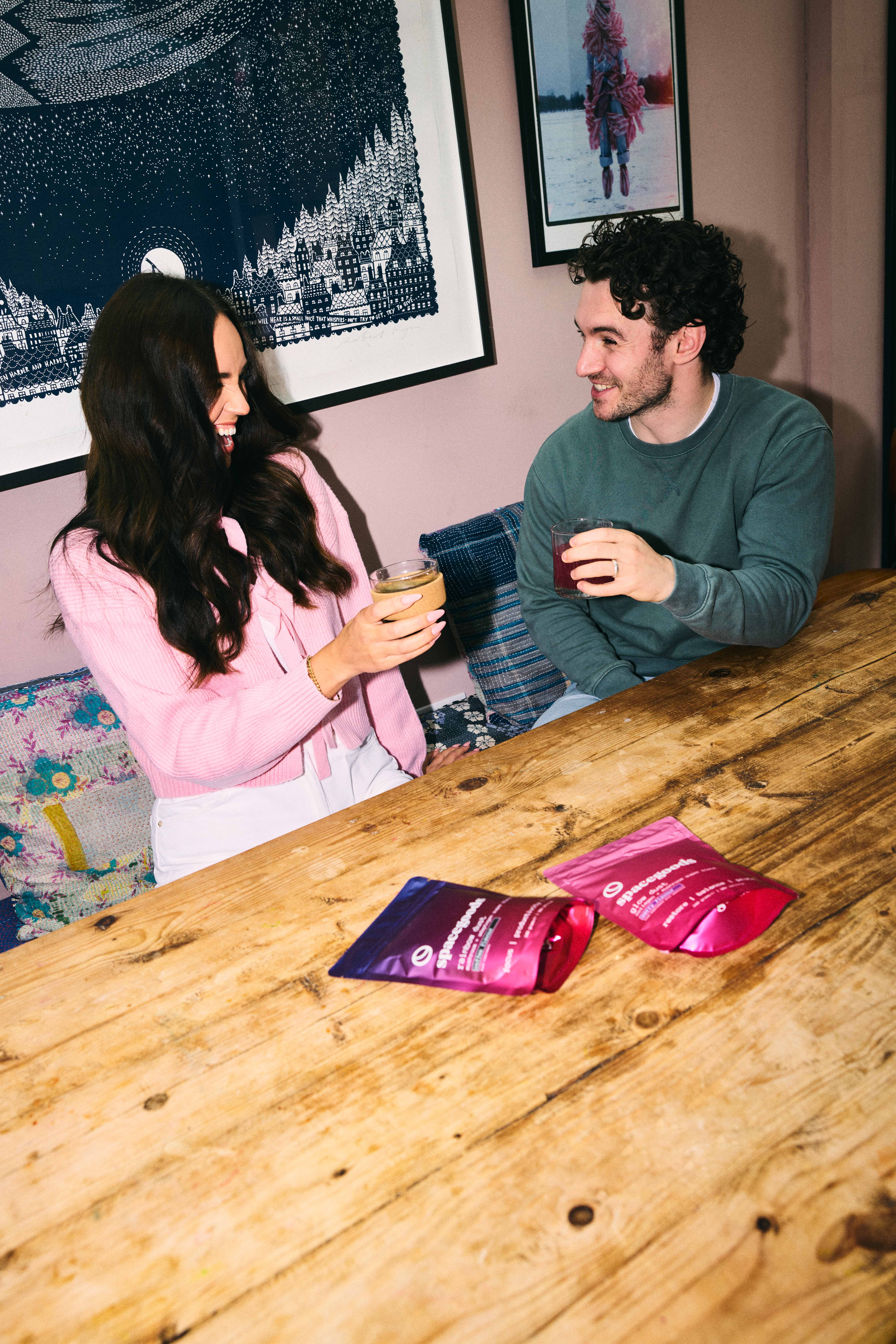 Two people smiling and holding drinks at a wooden table.