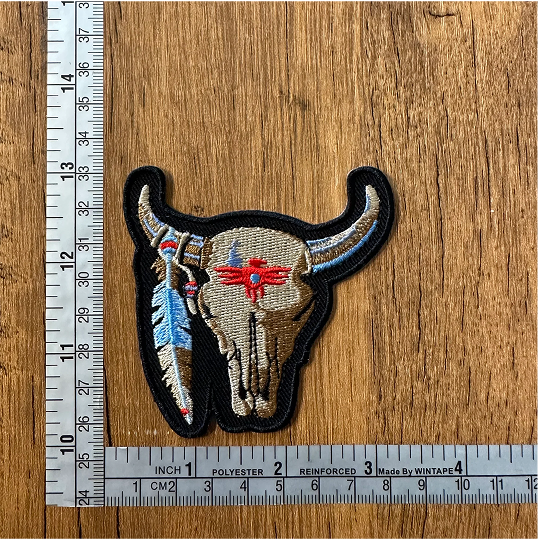 Embroidered bull skull patch with feather, measured by rulers on wooden surface.