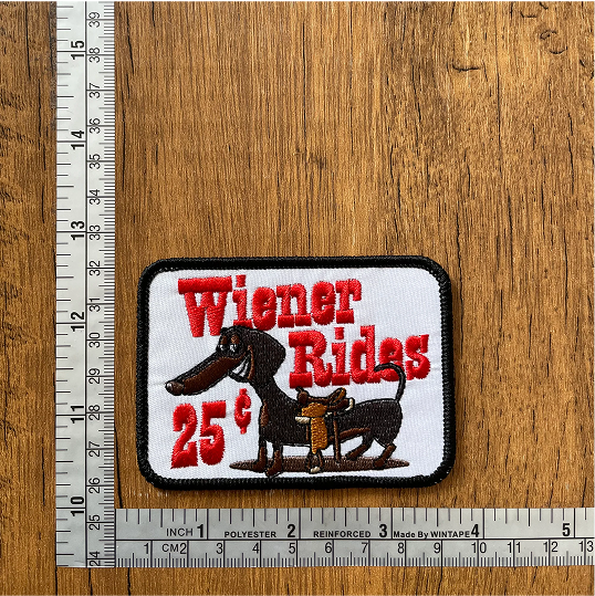 Embroidered patch with dachshund, text 'Wiener Rides 25¢,' on wooden surface with rulers.