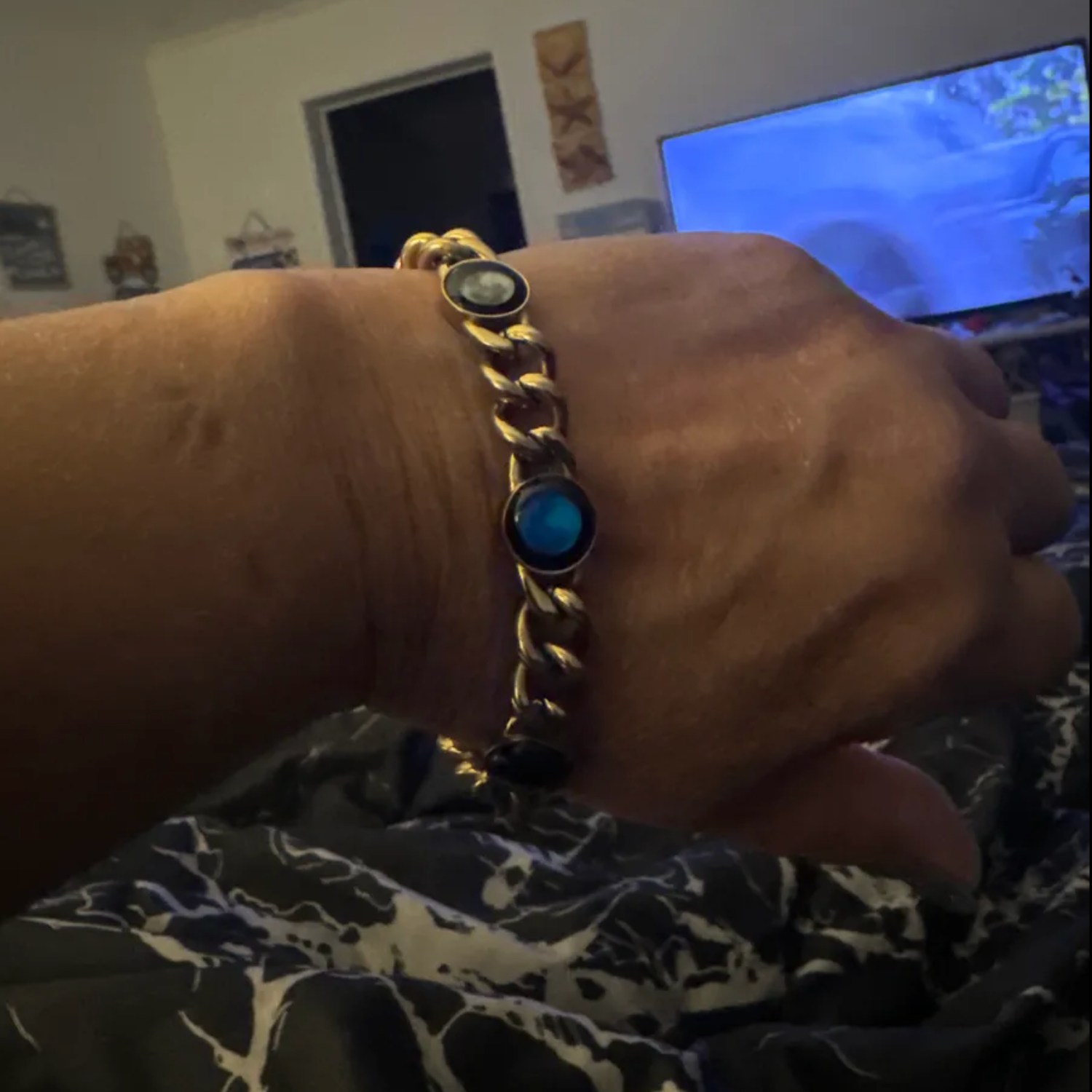 A hand wearing a gold bracelet with blue stones, in a room with a TV.