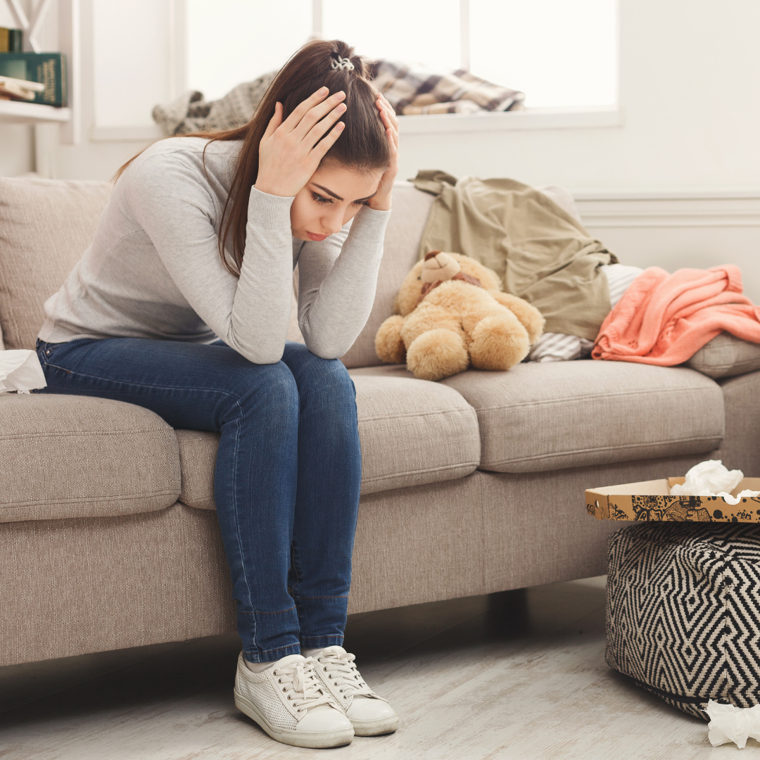 Woman sitting on a couch holding her head, surrounded by clutter.