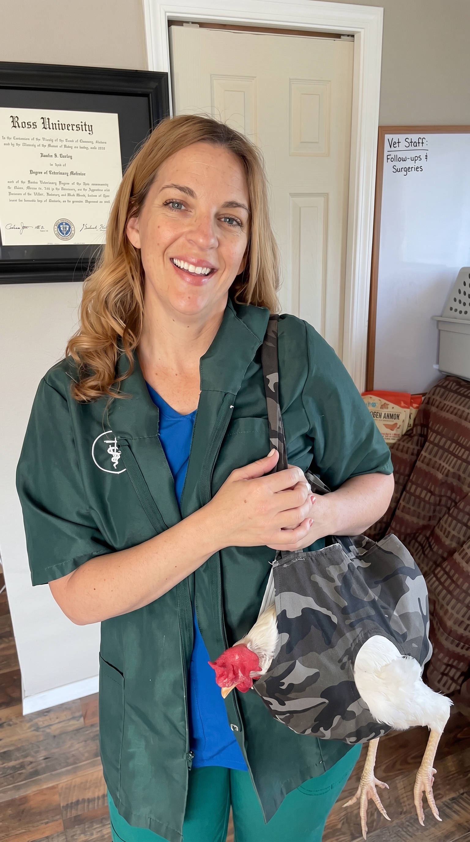 A woman in green scrubs smiles while holding a white chicken in a camouflage sling bag.