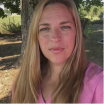 A woman with long blonde hair wearing a pink shirt stands outside in front of a tree.