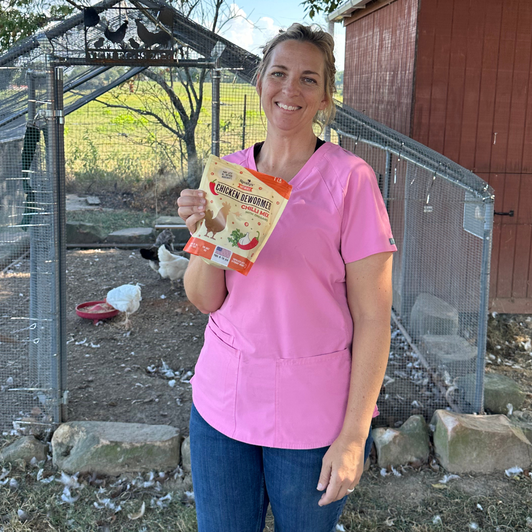 Person in pink shirt holding chicken dewormer package near a chicken coop.