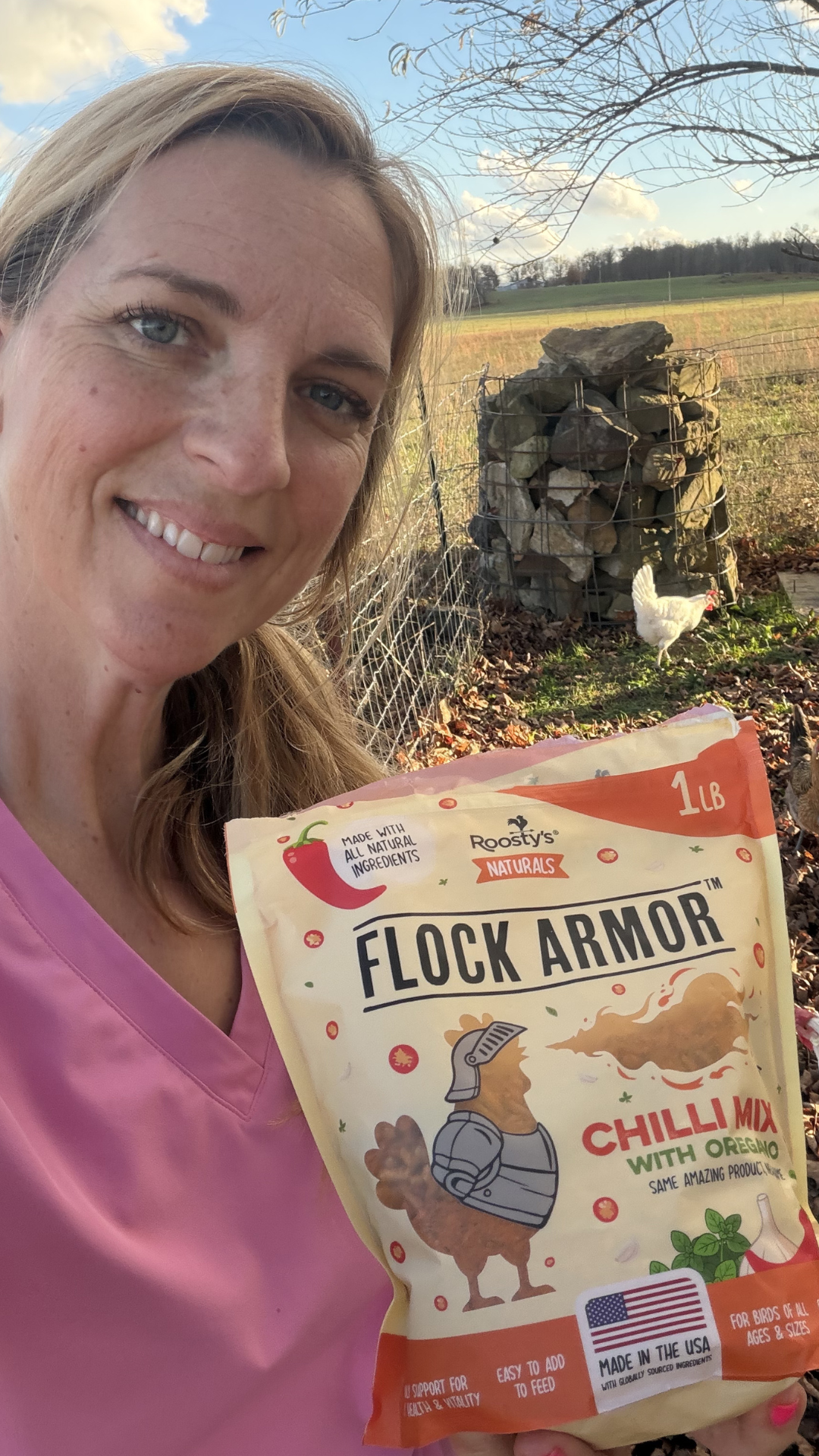 A woman smiles holding a bag of Flock Armor chicken feed with a chicken in the background.