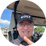A smiling man wearing a golf hat and glasses gestures from a golf cart.