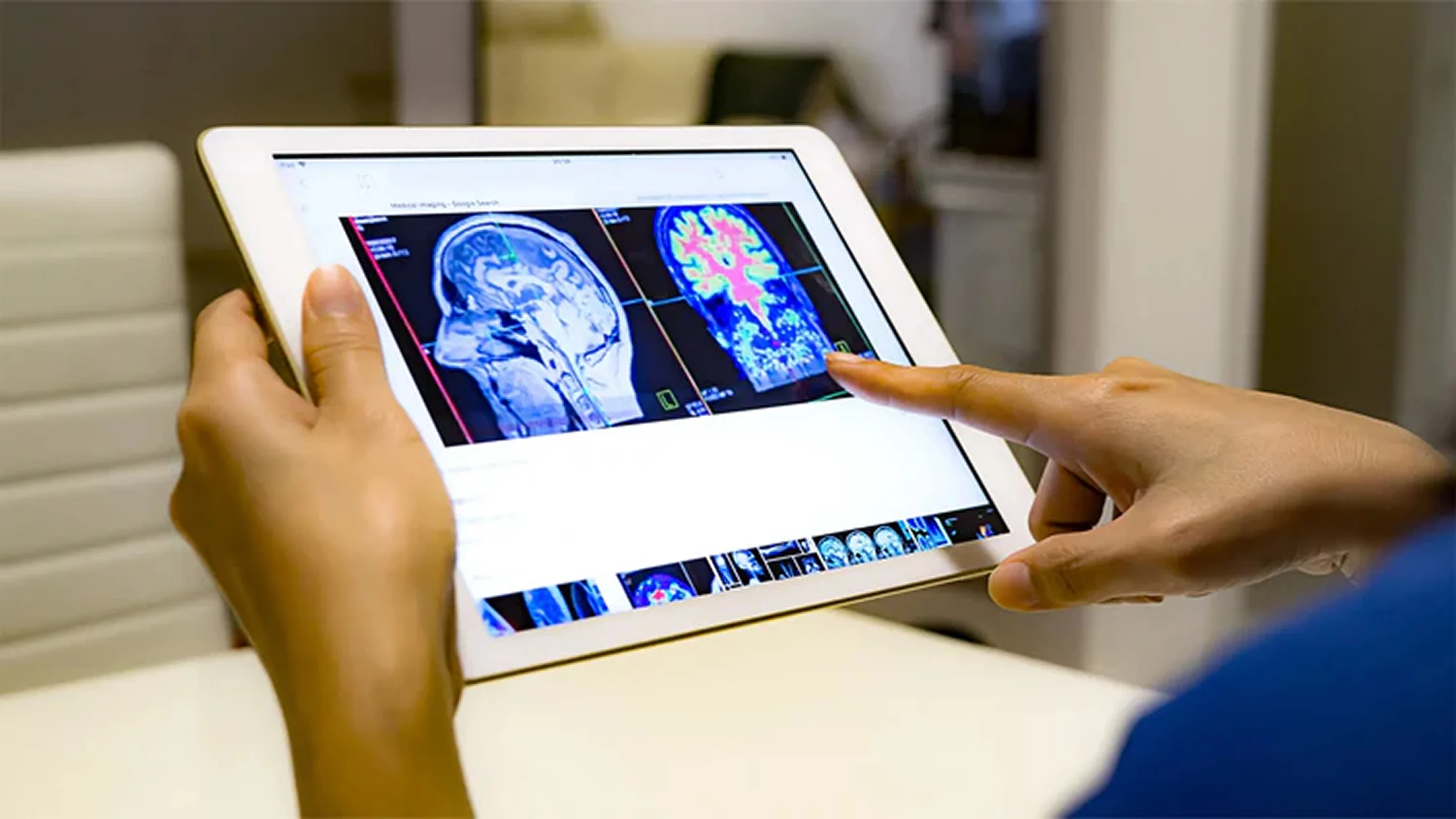 A person holds a white tablet and points to two colorful MRI brain scans on the screen.