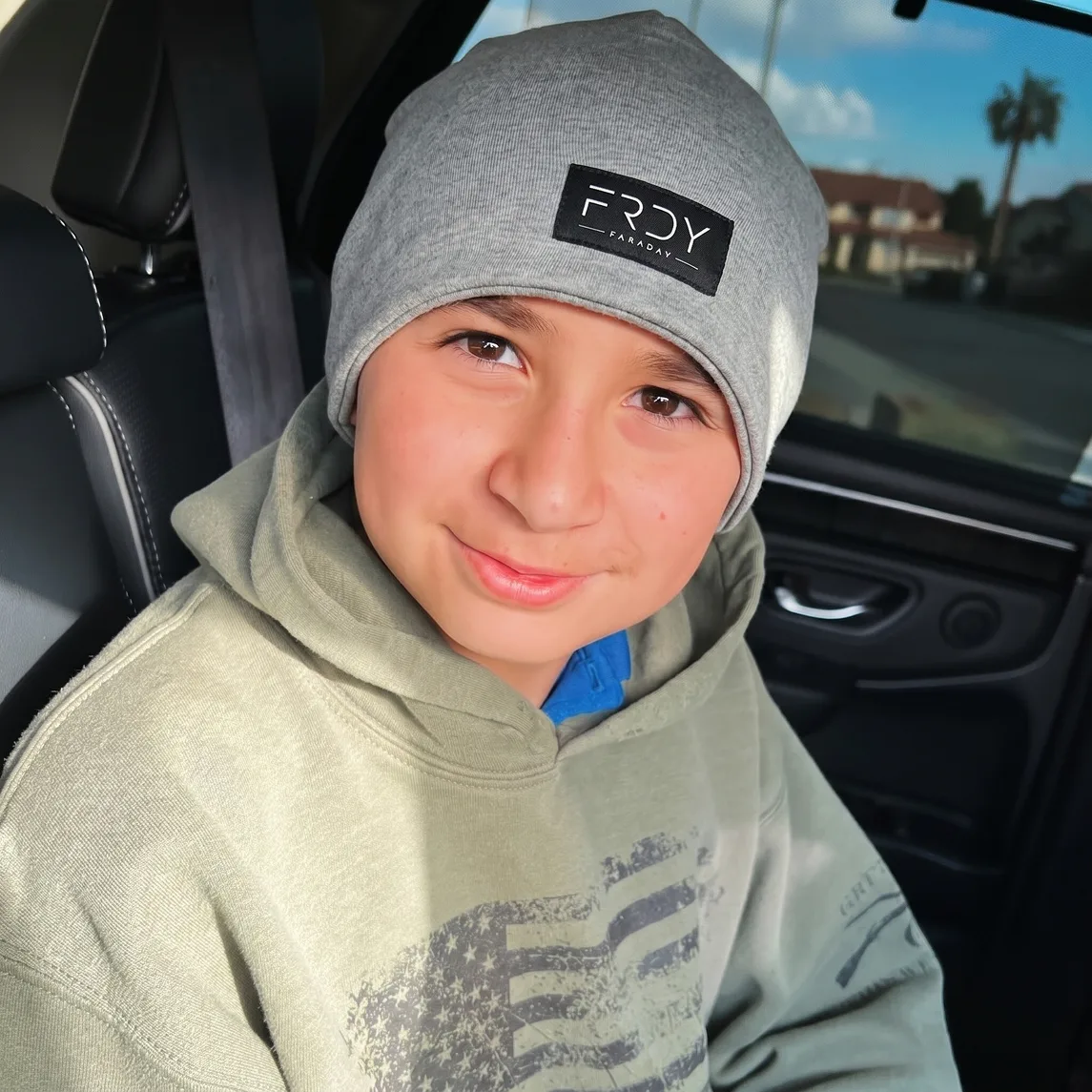A young boy in a grey beanie and a green hoodie smiles while sitting in a car.