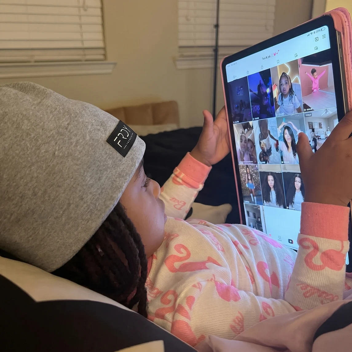 A young child in pajamas and a beanie lies down while holding a tablet and watching videos.