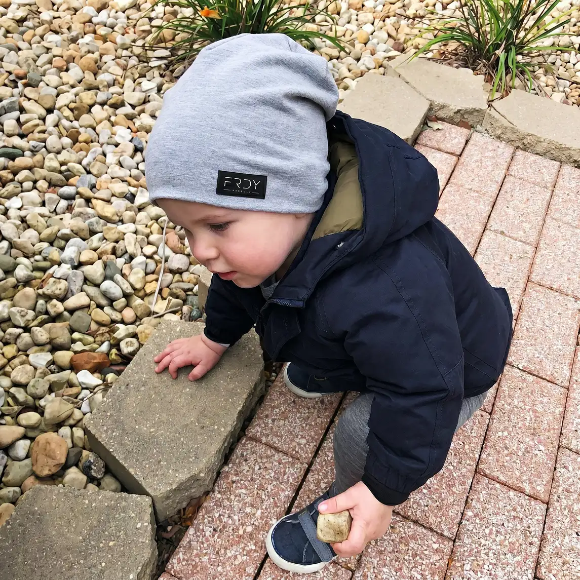 A young child in a grey beanie and blue jacket kneels on a path and plays with rocks.