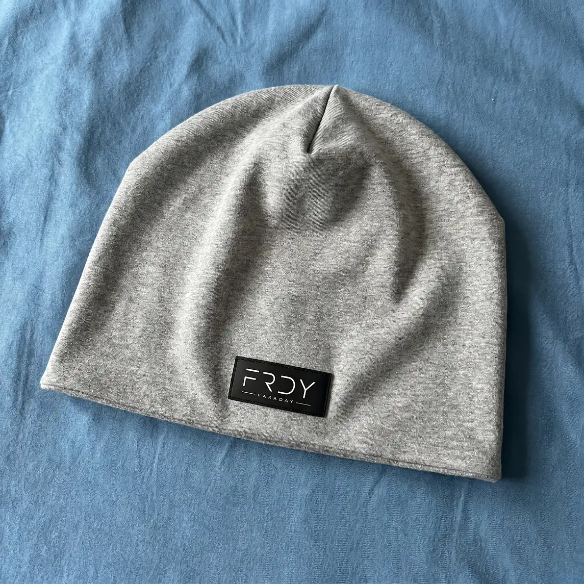 A gray beanie hat with a black 'FRDY FARADAY' label, resting on a blue fabric surface.