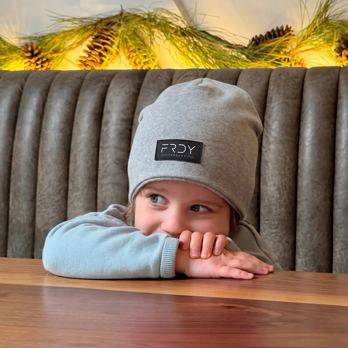 A young child in a grey beanie rests their chin on their hands at a wooden table.
