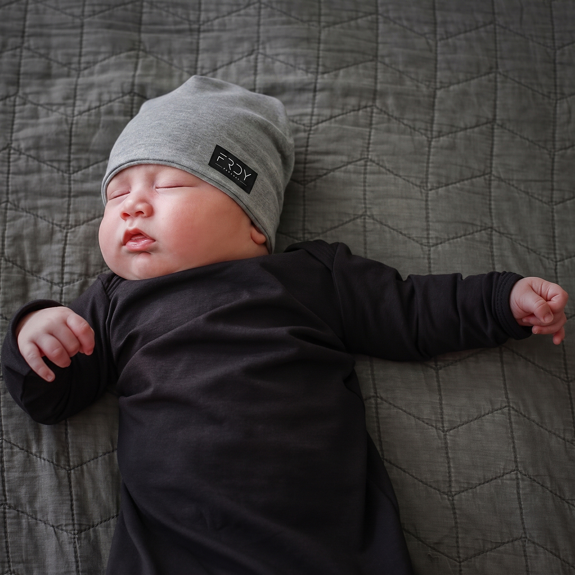 A baby wearing a grey beanie and a black outfit sleeps on a grey quilted blanket.