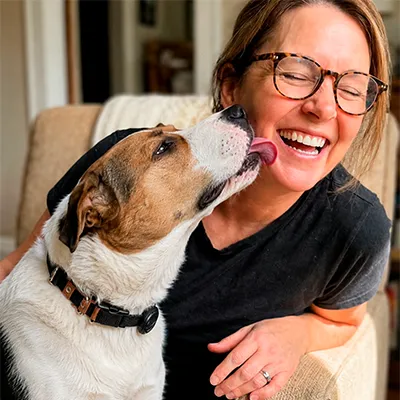 A dog licks the cheek of a laughing woman wearing glasses.
