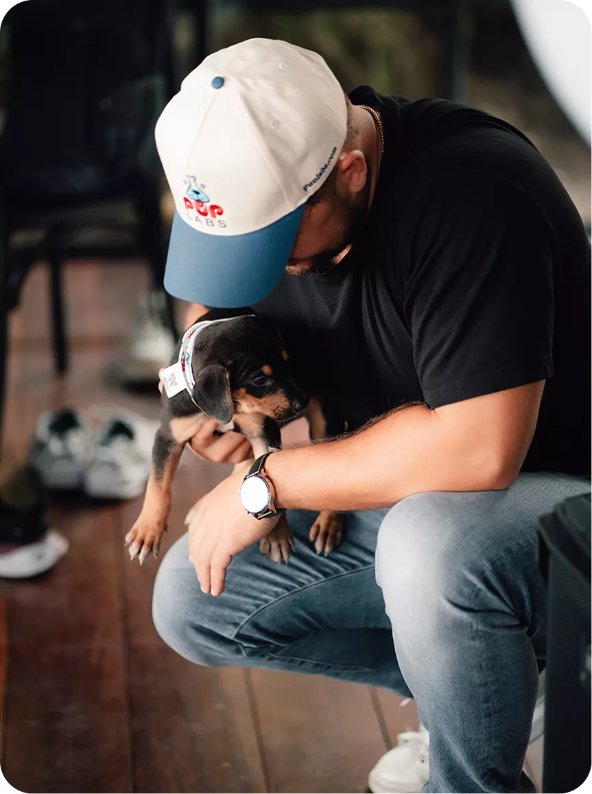 A man wearing a baseball cap squats down while holding a small black and tan puppy.
