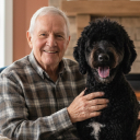 An older man in a plaid shirt smiles with his arm around a large, black, curly-haired dog.