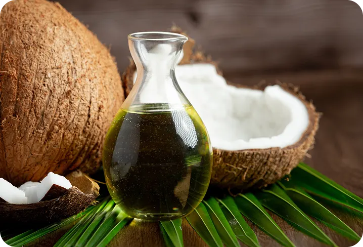 A glass decanter of coconut oil surrounded by fresh coconuts and palm leaves on a wooden surface.