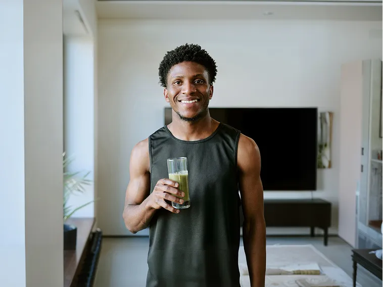 A smiling man in a black tank top holds a glass with a green smoothie in a living room.