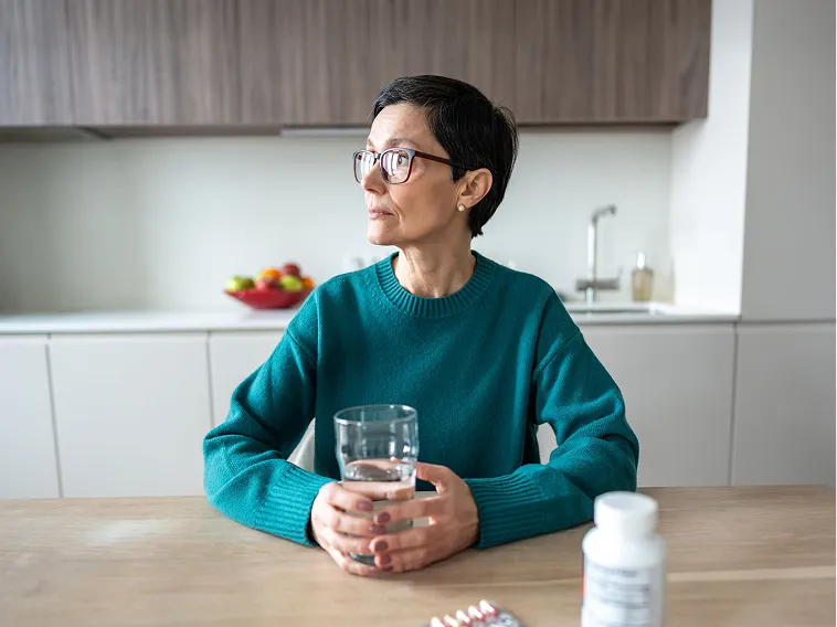 A woman with short hair and glasses sits at a kitchen table holding a glass of water.
