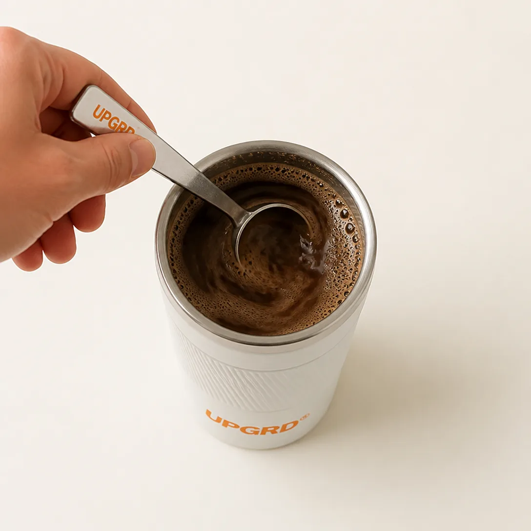A person's hand stirs a dark, frothy beverage in a white insulated tumbler with a silver spoon.